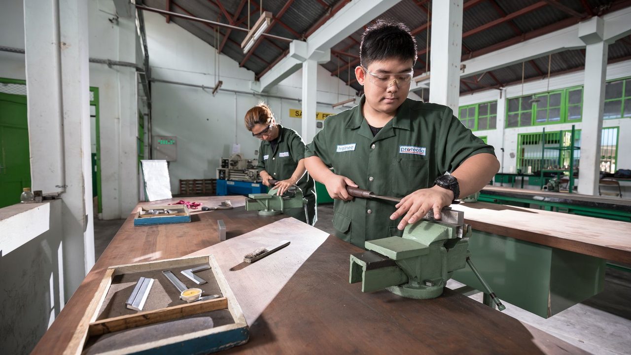 Two workers in a well-lit Indonesian workshop using clamping tools and safety gear for metalwork.