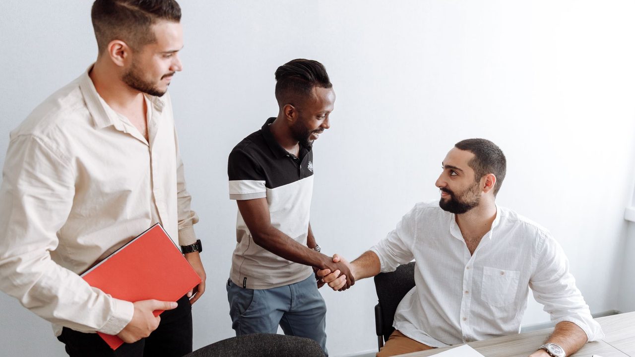 Three men in an office setting, shaking hands, representing teamwork and diversity.